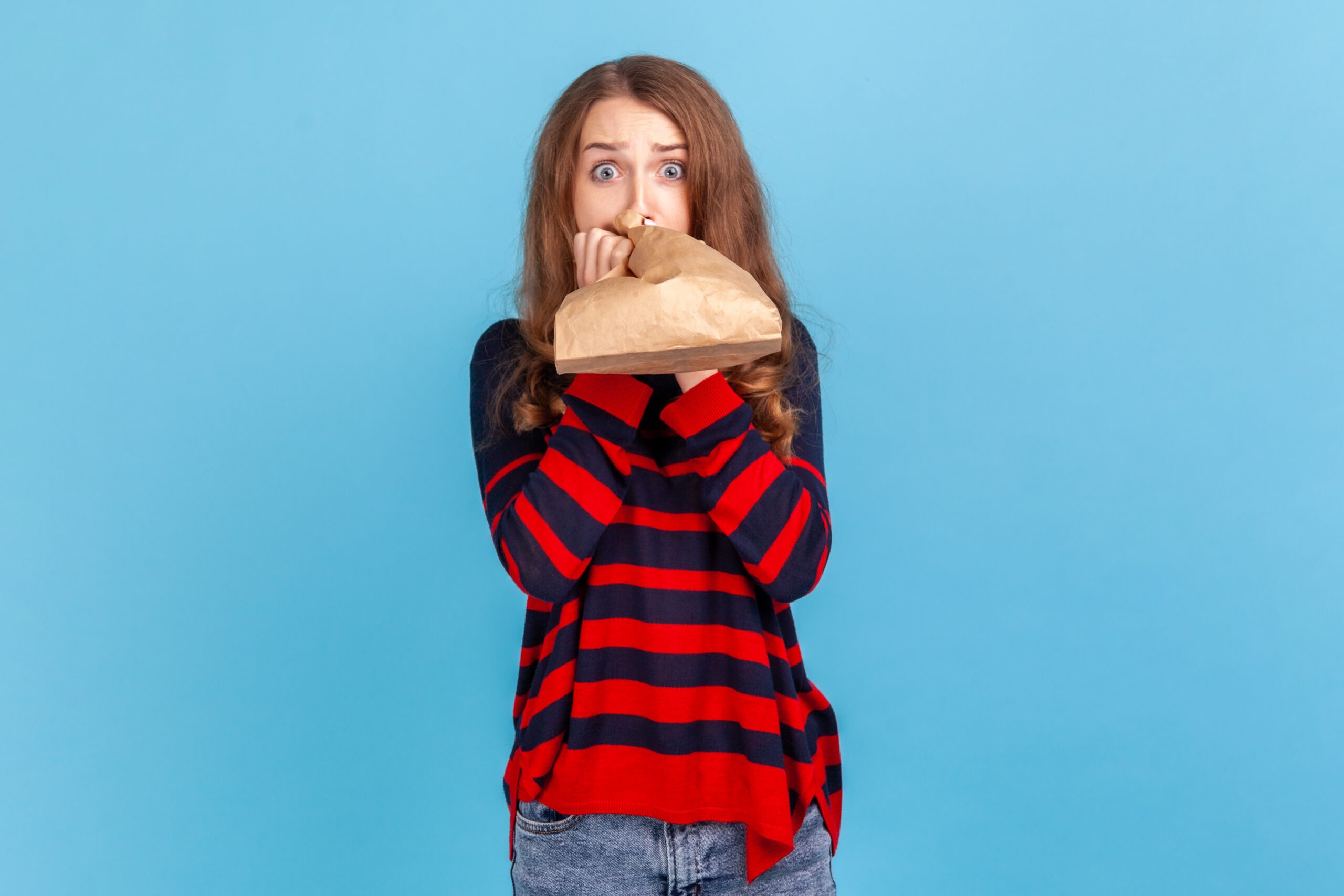 Woman breathing into paper bag to improve well-being, overcoming stress, looking at camera with fear