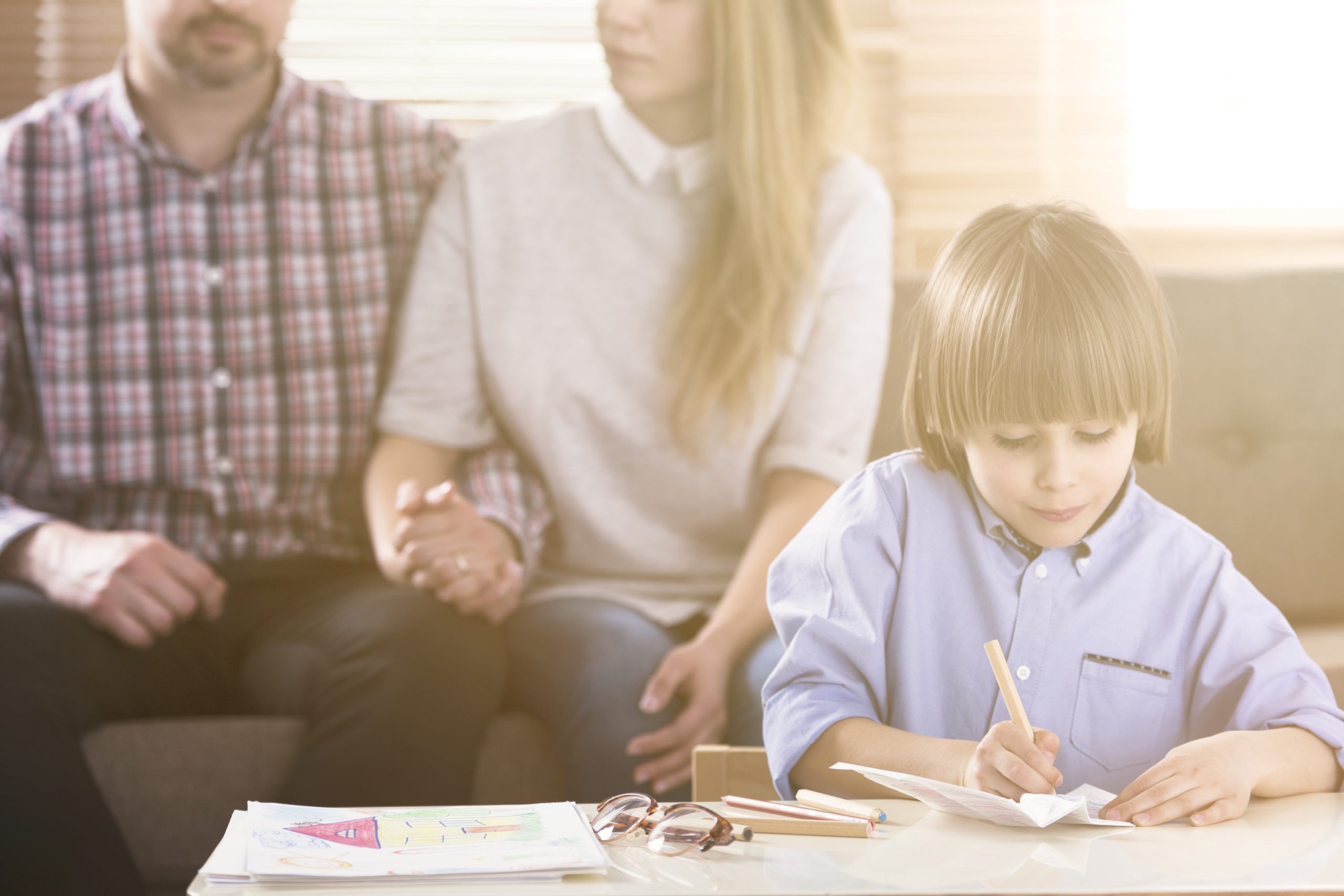 Man and woman holding hands on a couch and a boy in front drawing pictures by a table during a family psychotherapy session. Front view. Flare. Blurred background.